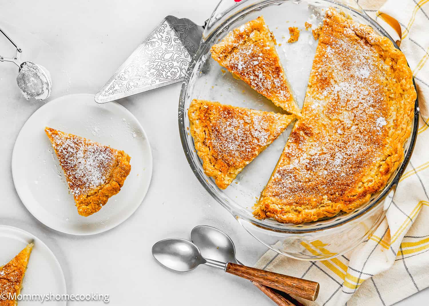 Overhead view of an Eggless Classic Chess Pie Recipe in a glass dish, with one slice served on a white plate beside two spoons and a yellow checkered kitchen towel.