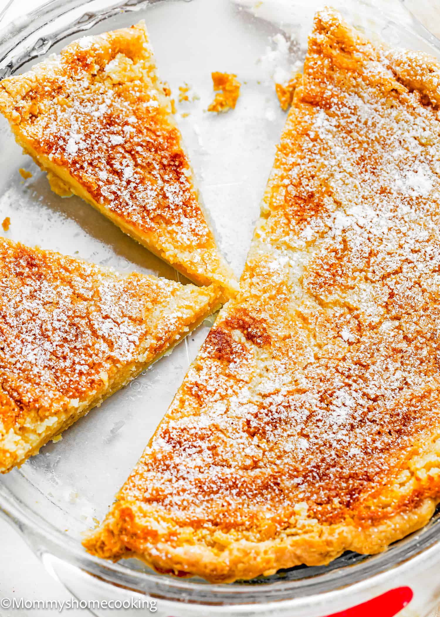 A partially eaten eggless classic chess pie in a glass pie dish, with three remaining slices topped with powdered sugar.