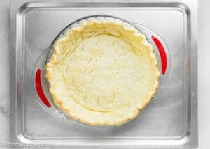 A baked pie crust with fork holes, perfect for an Eggless Classic Chess Pie Recipe, sits in a glass dish on a metal baking sheet.