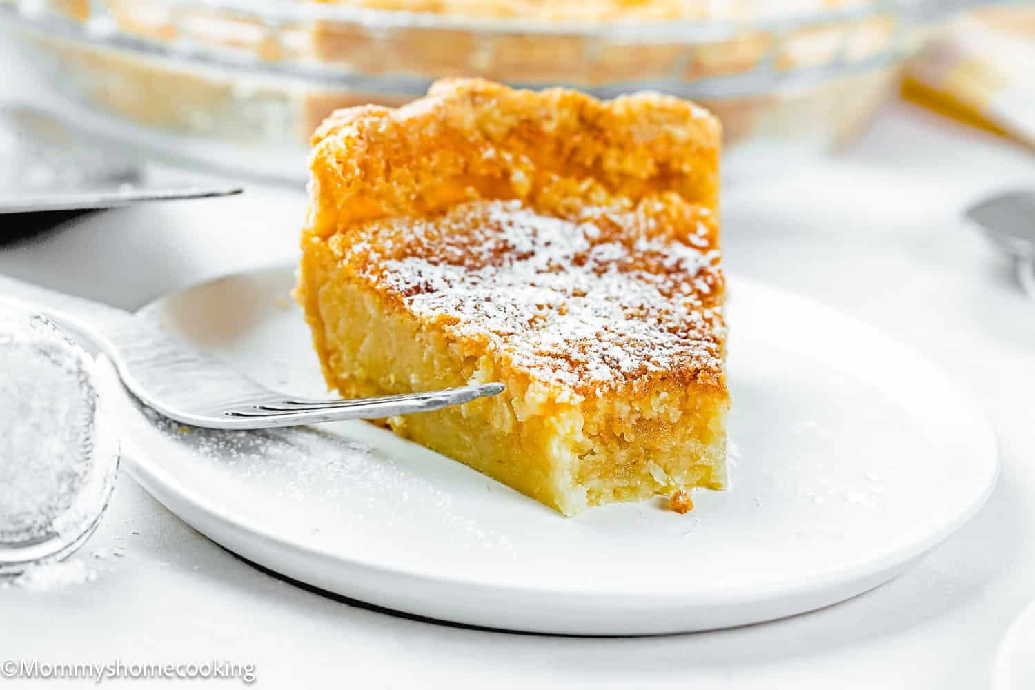 A slice of custard pie from an Eggless Classic Chess Pie Recipe, topped with powdered sugar on a white plate, with a fork beside it and a pie dish in the background.