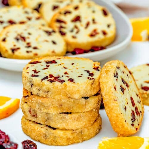 A stack of Eggless Cranberry Orange Icebox Cookies, coated in sugar, sits on a white surface next to orange slices, with more cookies on a plate in the background.