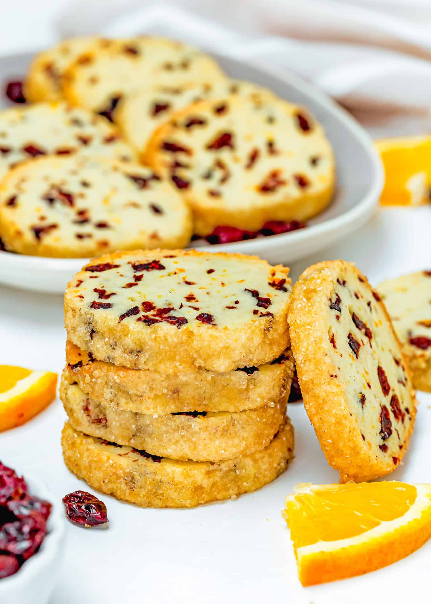 A stack of Eggless Cranberry Orange Icebox Cookies, coated in sugar, sits on a white surface next to orange slices, with more cookies on a plate in the background.