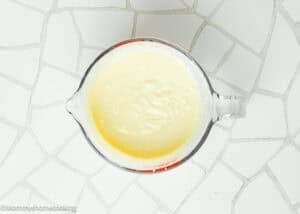 A glass measuring cup filled with thick, pale yellow condensed milk—perfect for creating an Eggless Creamy Lemon Pie—sits on a white tiled surface.