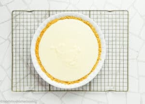 An Eggless Creamy Lemon Pie with a graham cracker crust and smooth, pale filling sits on a round white dish atop a cooling rack on a white tiled surface.