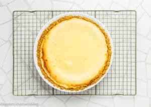 A baked cheesecake with a golden graham cracker crust sits on a cooling rack over a white tiled surface, reminiscent of an Eggless Creamy Lemon Pie in its smooth, luscious texture.