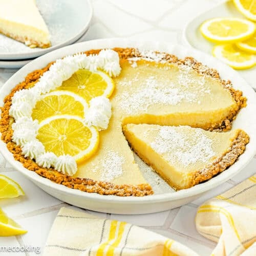An Eggless Creamy Lemon Pie with a graham cracker crust is garnished with whipped cream, lemon slices, and powdered sugar. One slice is cut and partially removed from the pie.