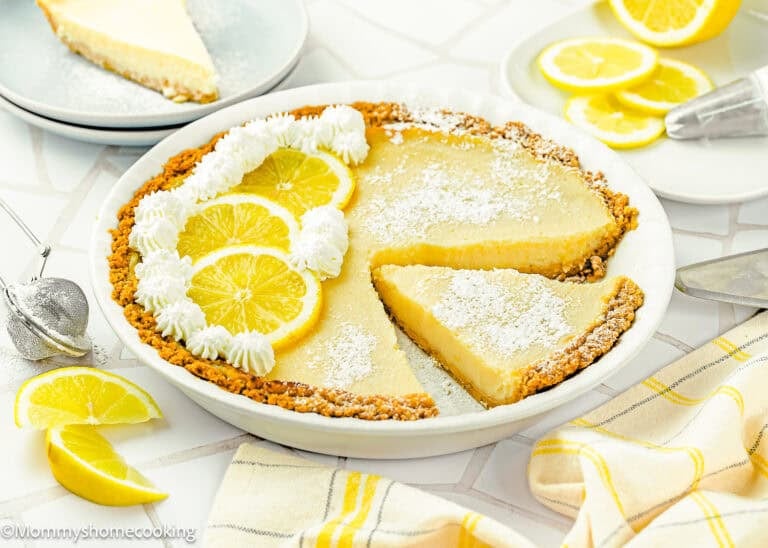 An Eggless Creamy Lemon Pie with a graham cracker crust is garnished with whipped cream, lemon slices, and powdered sugar. One slice is cut and partially removed from the pie.