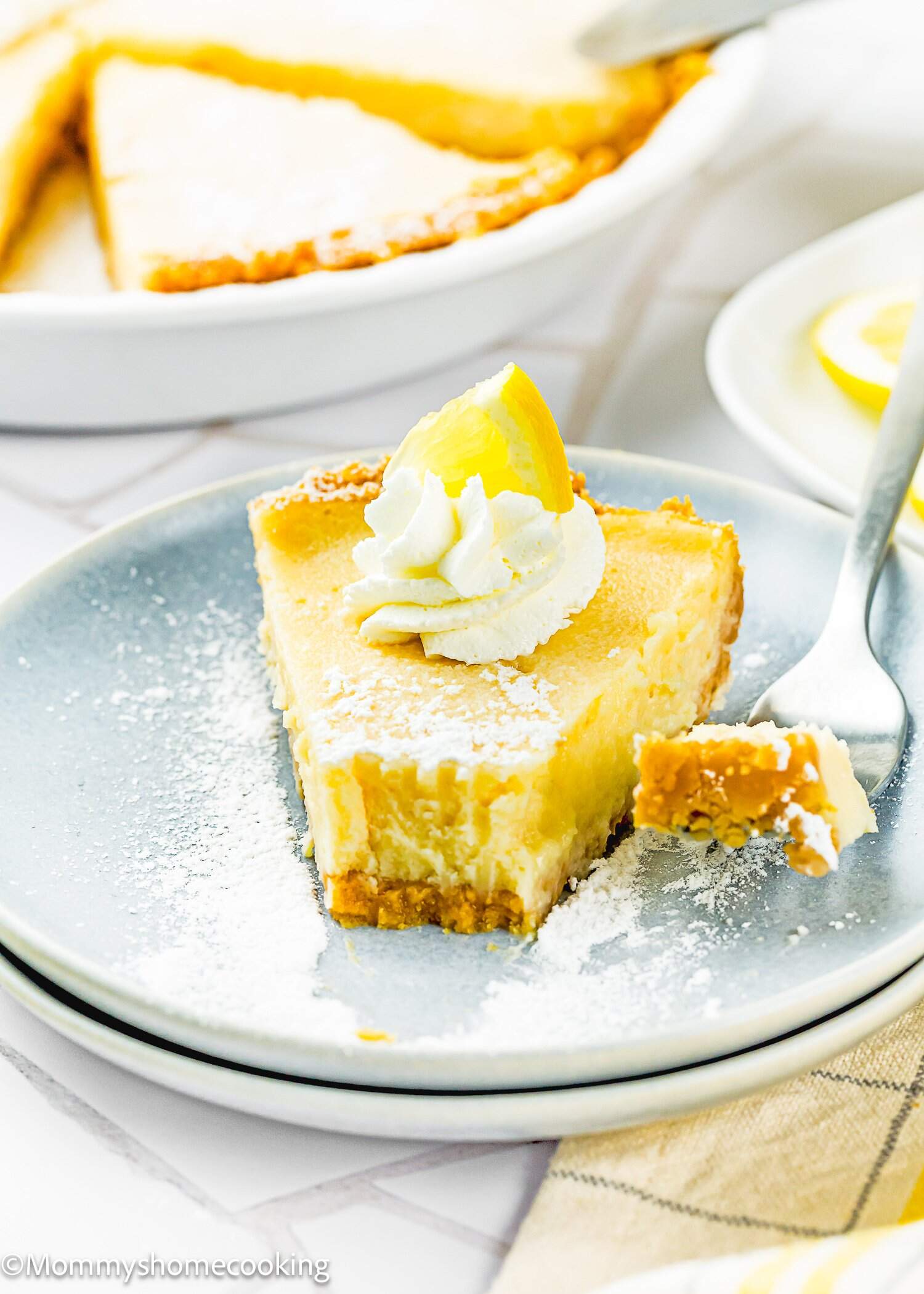 A slice of Eggless Creamy Lemon Pie topped with whipped cream and a lemon wedge sits on a plate, with a fork holding a bite-sized piece.