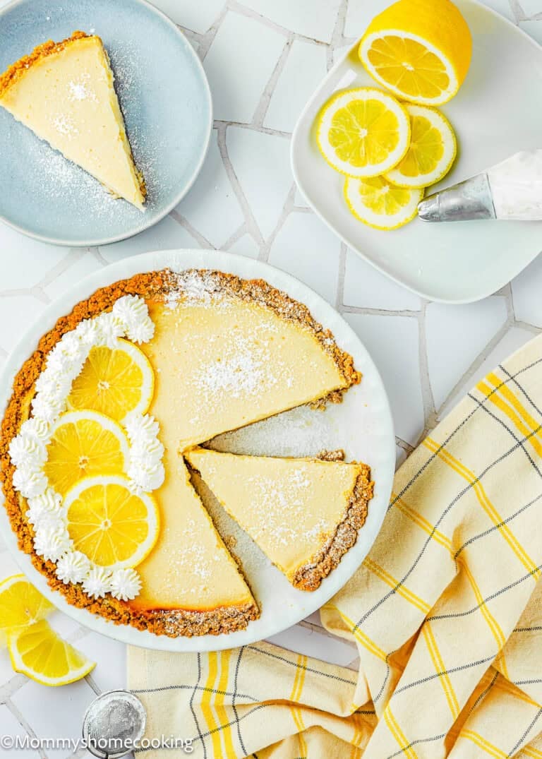 A slice of Eggless Creamy Lemon Pie is served on a plate, garnished with lemon slices, whipped cream, and powdered sugar. Nearby are a sliced lemon and a piping bag on a tiled surface.