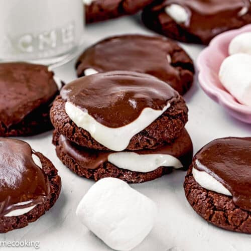 Eggless Hot Chocolate Cookies topped with toasted marshmallow and a layer of chocolate ganache, arranged beside marshmallows and a glass of milk.