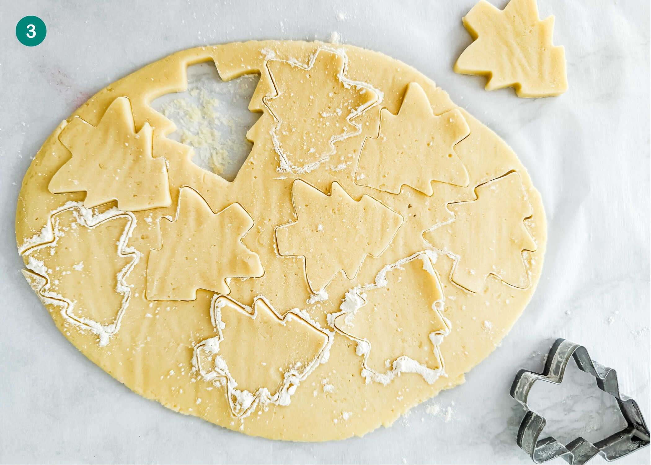 Rolled-out cookie dough with multiple Eggless Marble Christmas Tree Cookies cut out, a tree-shaped cookie cutter, and flour sprinkled on top.