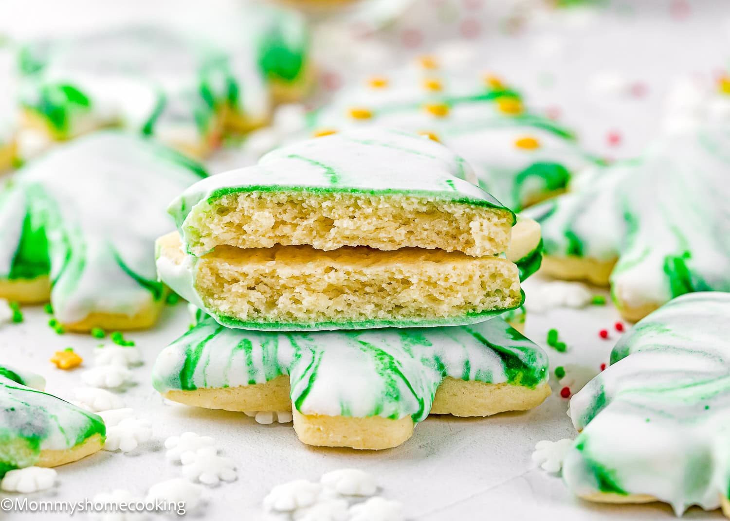 A stack of Eggless Marble Christmas Tree Cookies shaped like shamrocks, with green and white marbled icing, one cookie broken in half to reveal the soft interior.