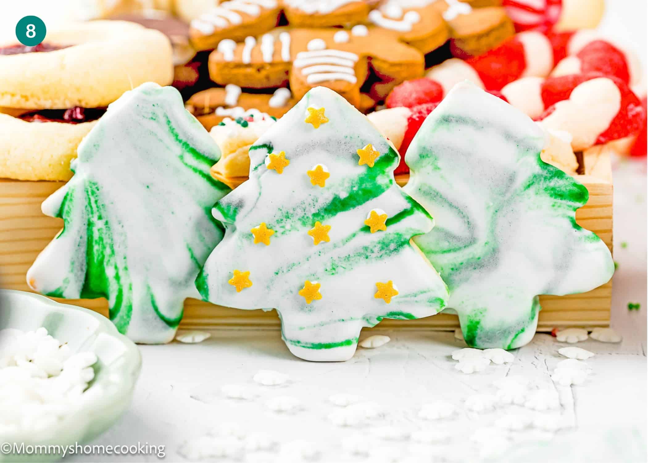 Three Eggless Marble Christmas Tree Cookies with green and white icing, one decorated with yellow star sprinkles, displayed among other assorted holiday cookies.