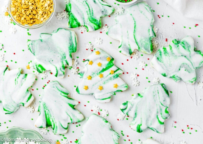 Eggless Marble Christmas Tree Cookies shaped like trees, decorated with white and green icing, and surrounded by red, green, and white sprinkles on a white surface.