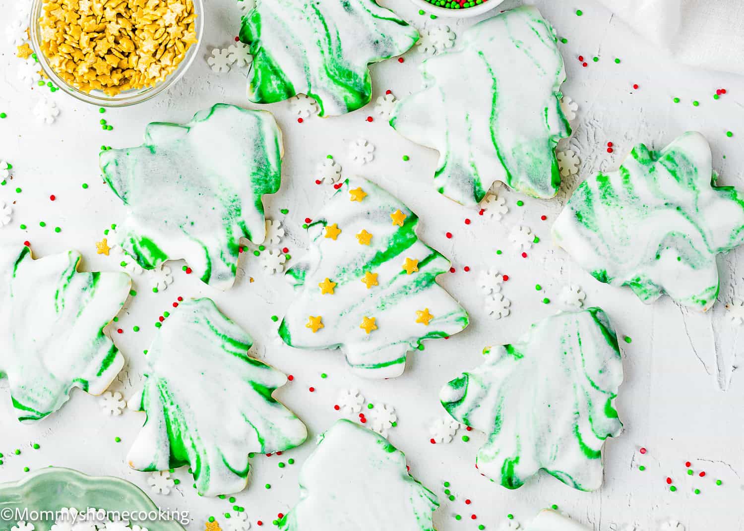 Eggless Marble Christmas Tree Cookies shaped like trees, decorated with white and green icing, and surrounded by red, green, and white sprinkles on a white surface.