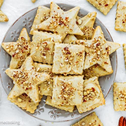A plate of Eggless Pecan Sugar Cookies with Brown Butter Icing in star and square shapes, topped with chopped pecans, surrounded by extra cookies and nuts on a white surface.