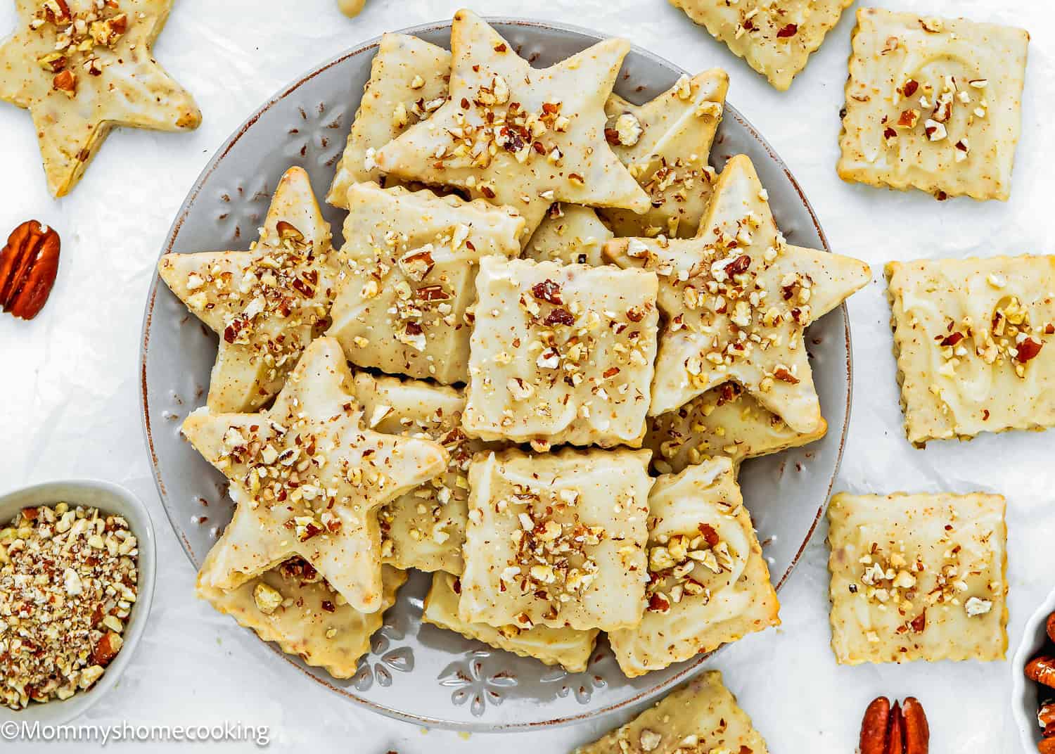A plate of Eggless Pecan Sugar Cookies with Brown Butter Icing in star and square shapes, topped with chopped pecans, surrounded by extra cookies and nuts on a white surface.