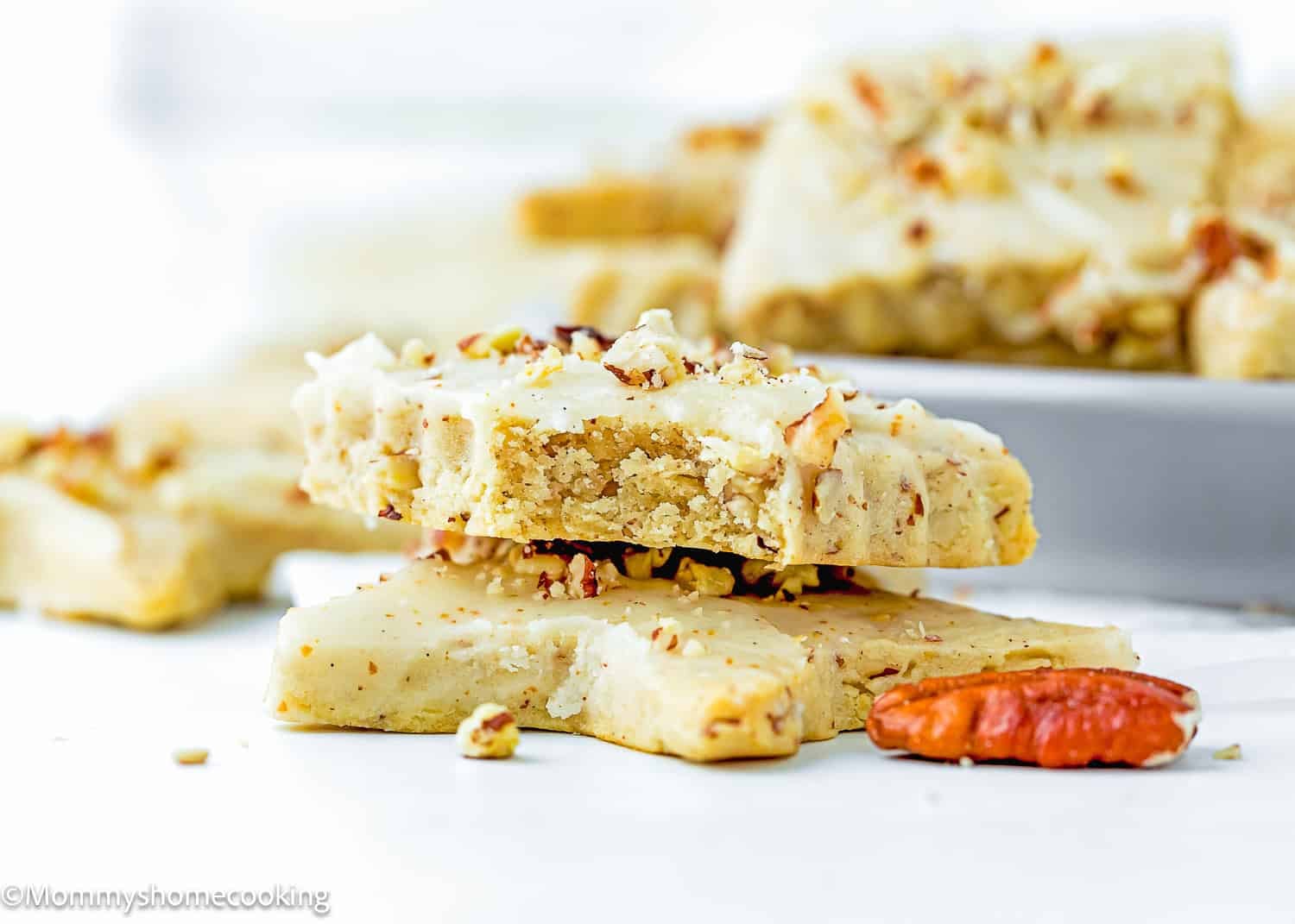 Two Eggless Pecan Sugar Cookies with Brown Butter Icing are stacked on a white surface, with more bars and pecans visible in the background.