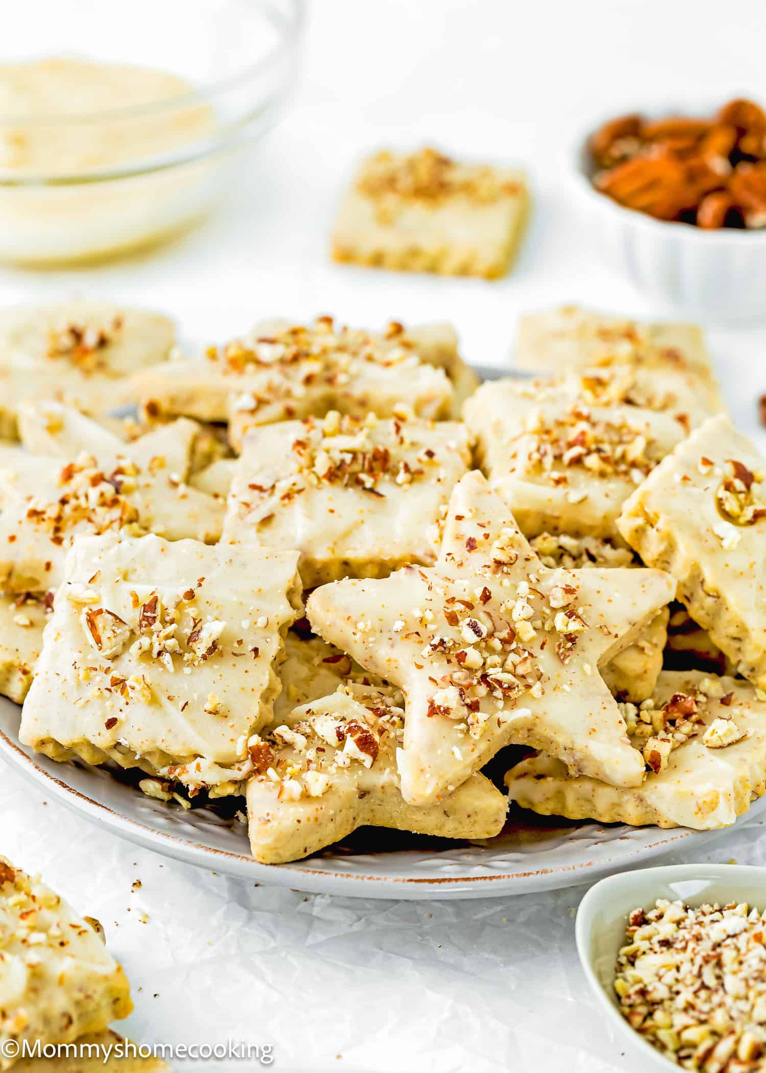 A plate of Eggless Pecan Sugar Cookies with Brown Butter Icing in star and square shapes, topped with chopped nuts, sits with bowls of ingredients in the background.