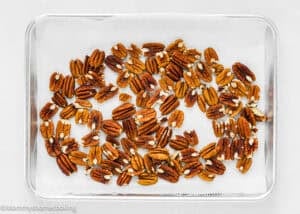 A glass tray lined with parchment paper holds a single layer of roasted pecan halves, ready for making Eggless Pecan Sugar Cookies with Brown Butter Icing.
