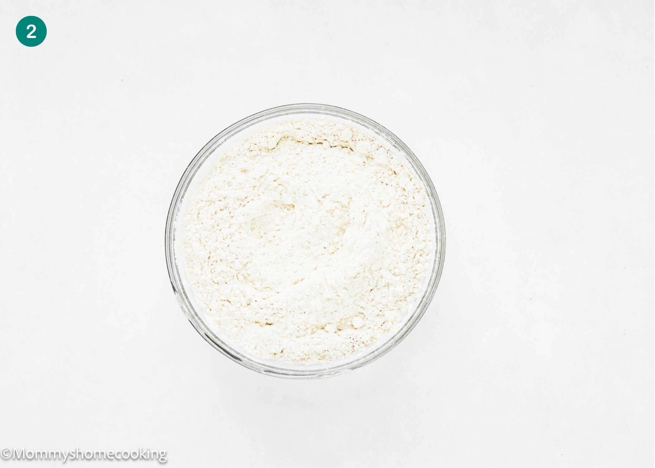 A clear glass bowl filled with white flour is placed on a plain white background, perfect for making Eggless Pecan Sugar Cookies with Brown Butter Icing.