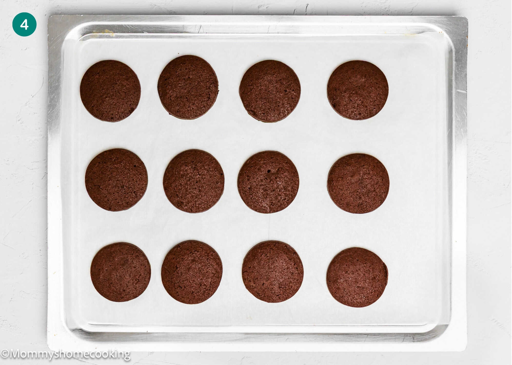 A baking tray lined with parchment paper holds twelve evenly spaced, round Eggless Peppermint Bark Cookies.