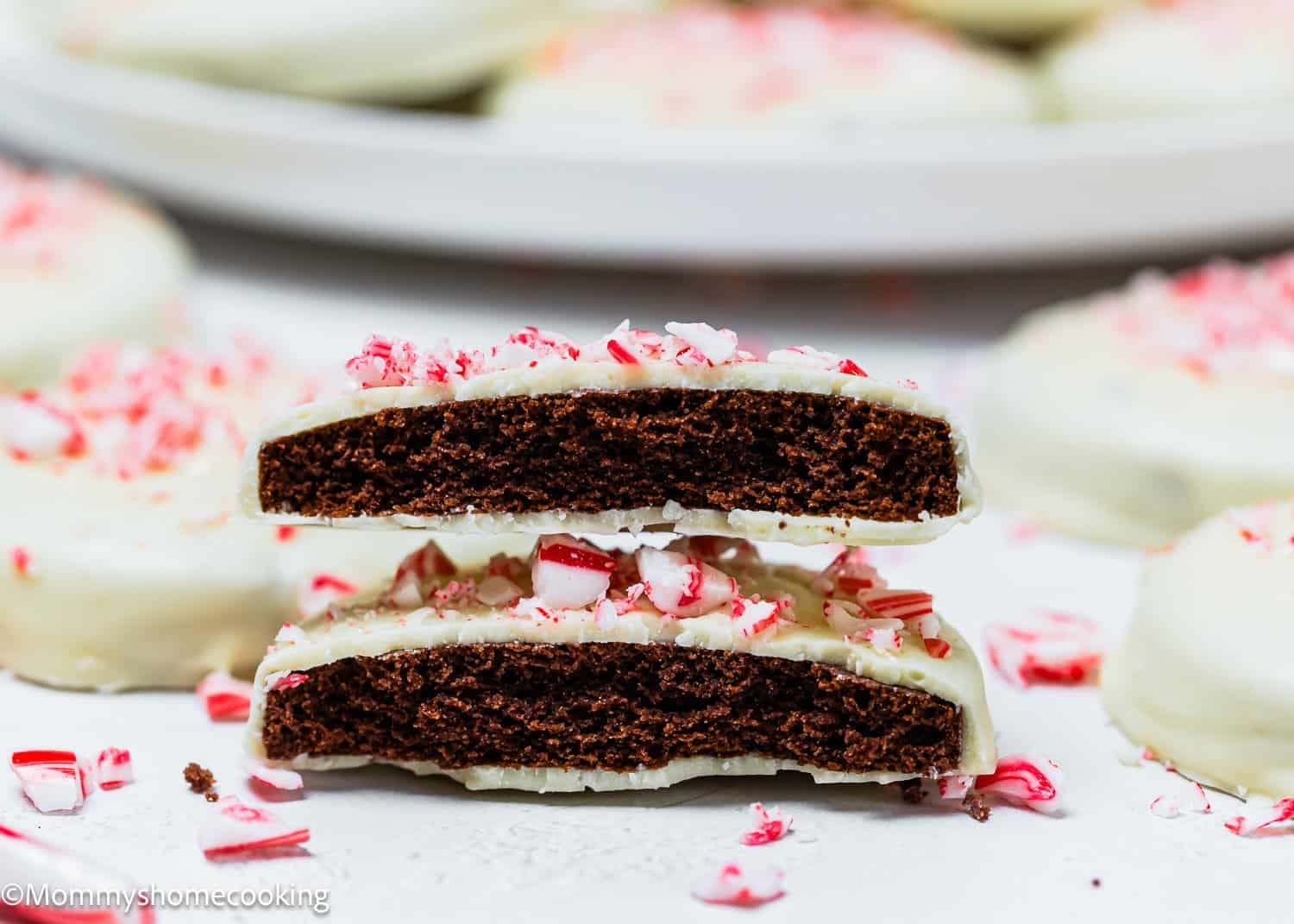 A close-up of two Eggless Peppermint Bark Cookies coated in white chocolate and crushed peppermint, stacked to reveal their inside; more cookies are blurred in the background.