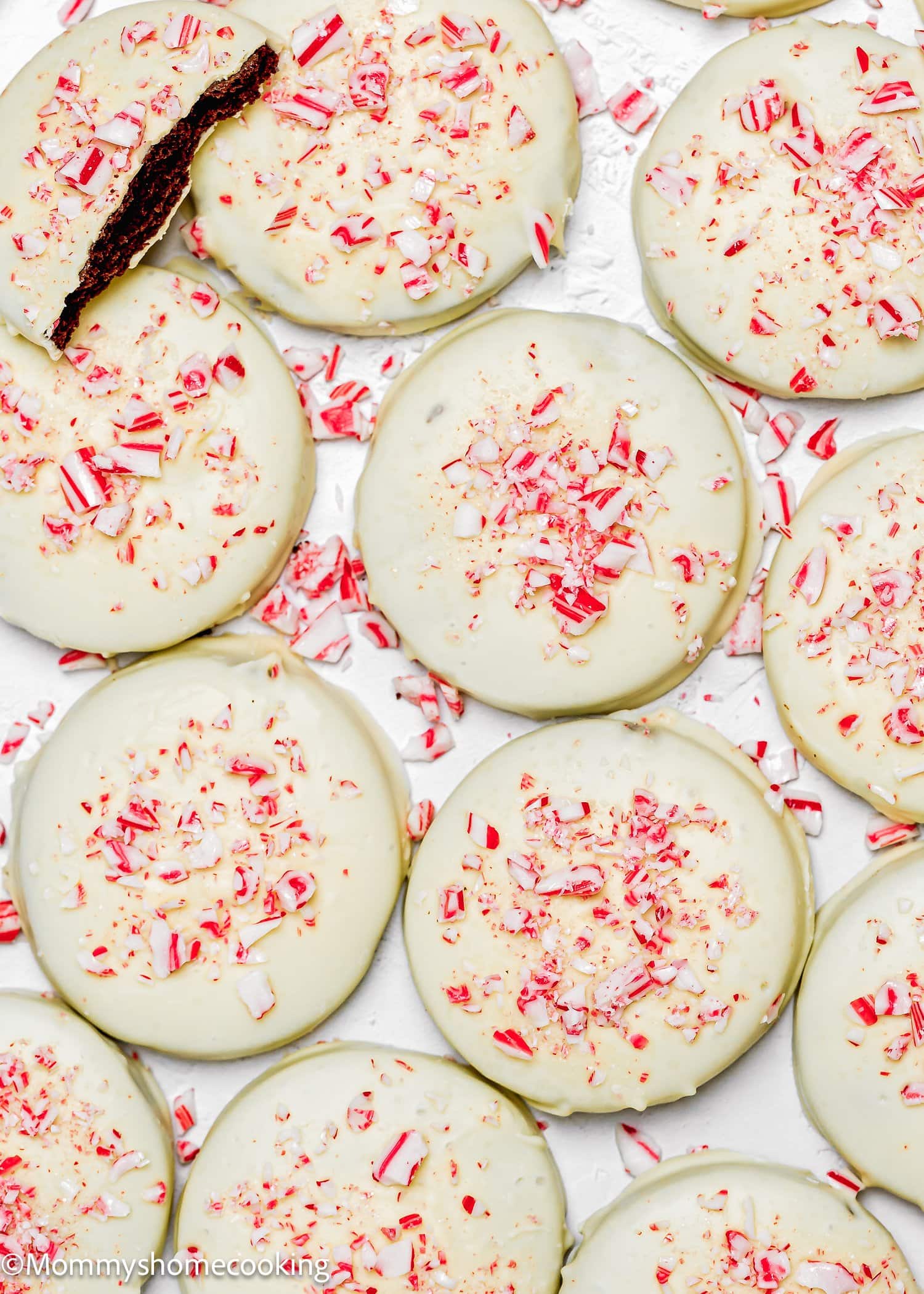 Eggless Peppermint Bark Cookies: Round chocolate cookies coated with white icing and sprinkled with crushed peppermint candies, arranged on a white surface. One cookie is partially bitten.