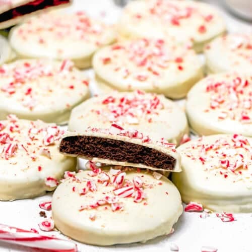 A batch of Eggless Peppermint Bark Cookies, round chocolate cookies coated in white icing and topped with crushed peppermint candy, with one cookie showing the chocolate interior.
