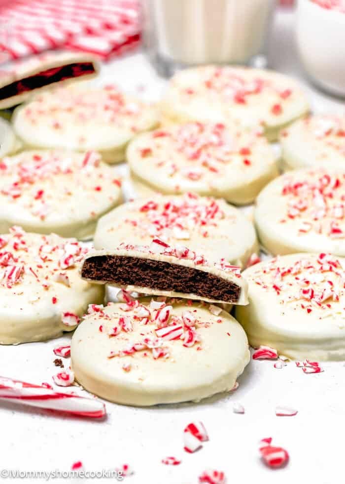 A batch of Eggless Peppermint Bark Cookies, round chocolate cookies coated in white icing and topped with crushed peppermint candy, with one cookie showing the chocolate interior.
