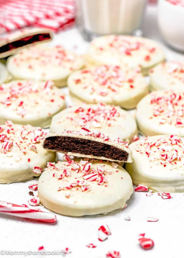 A batch of Eggless Peppermint Bark Cookies, round chocolate cookies coated in white icing and topped with crushed peppermint candy, with one cookie showing the chocolate interior.