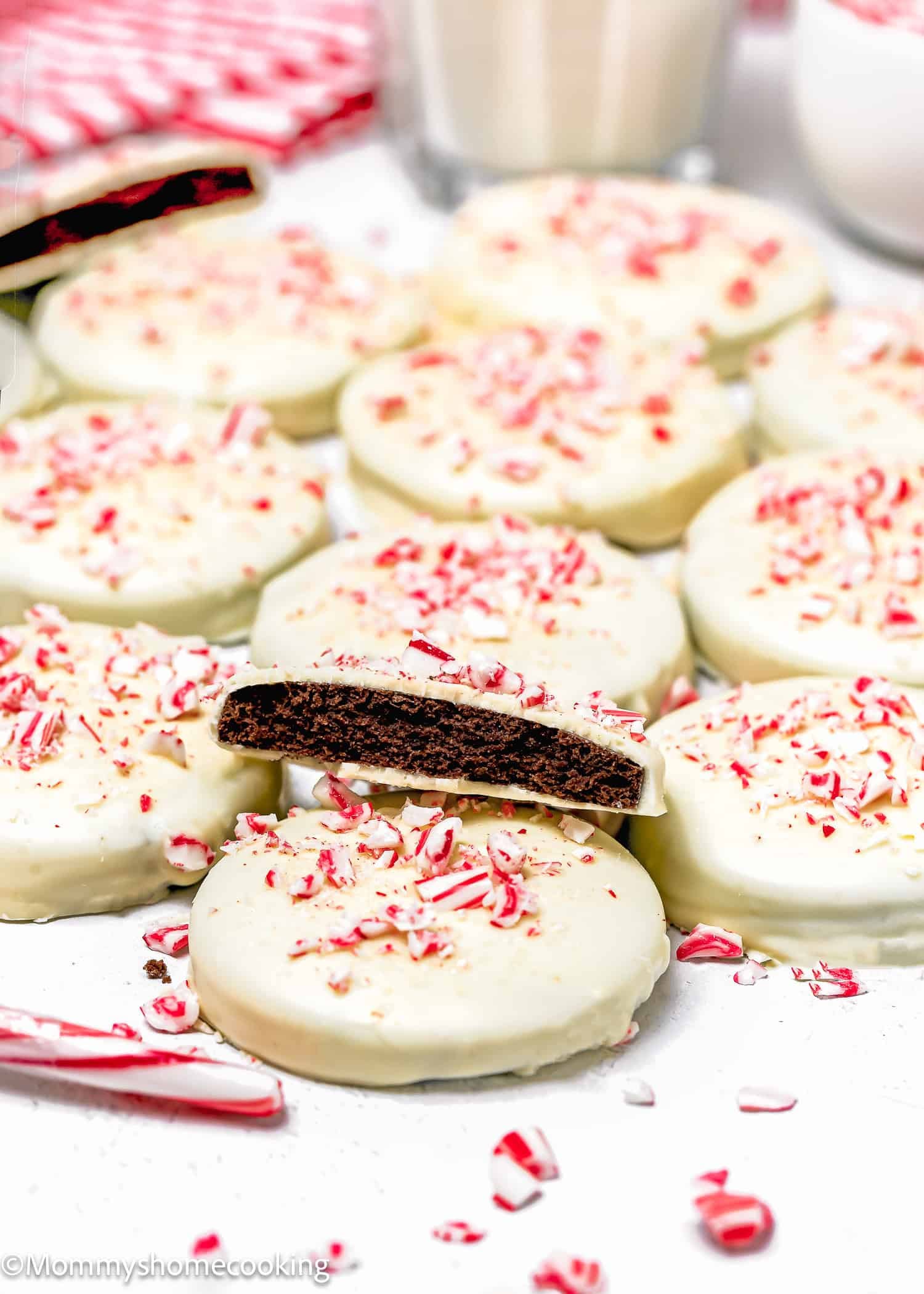 A batch of Eggless Peppermint Bark Cookies, round chocolate cookies coated in white icing and topped with crushed peppermint candy, with one cookie showing the chocolate interior.