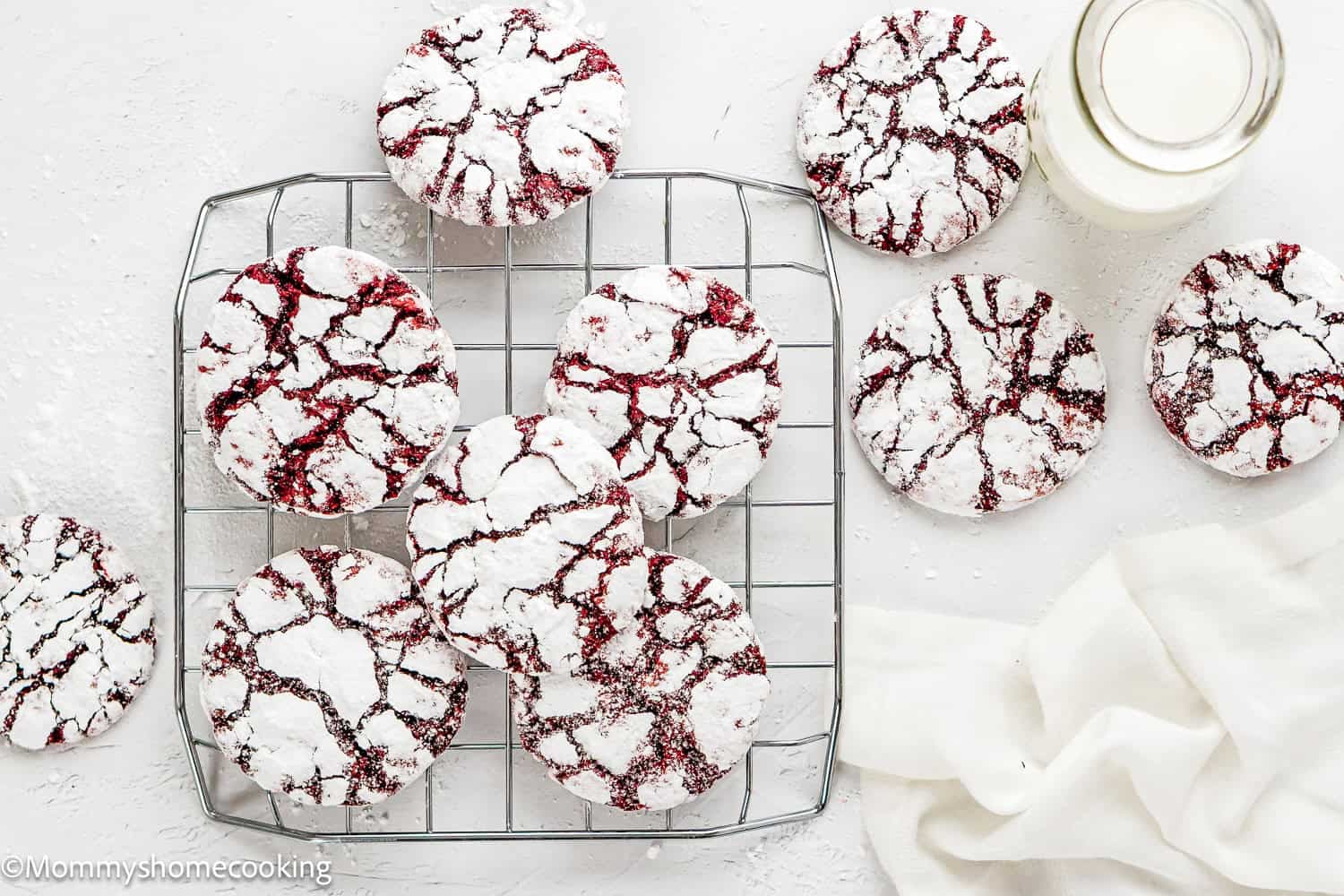 A cooling rack holds several Eggless Red Velvet Crinkle Cookies dusted with powdered sugar. More cookies, a glass of milk, and a white cloth are nearby on a light surface.