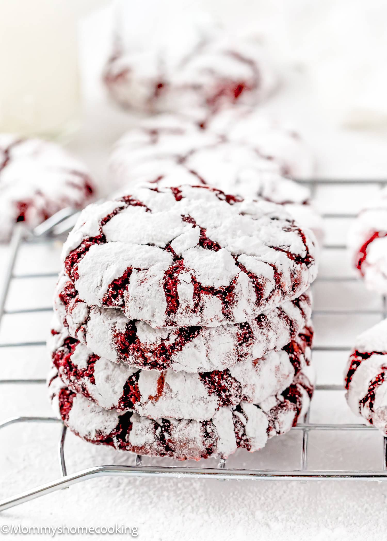A stack of three eggless red velvet crinkle cookies coated in powdered sugar sits on a cooling rack, with more cookies in the background.