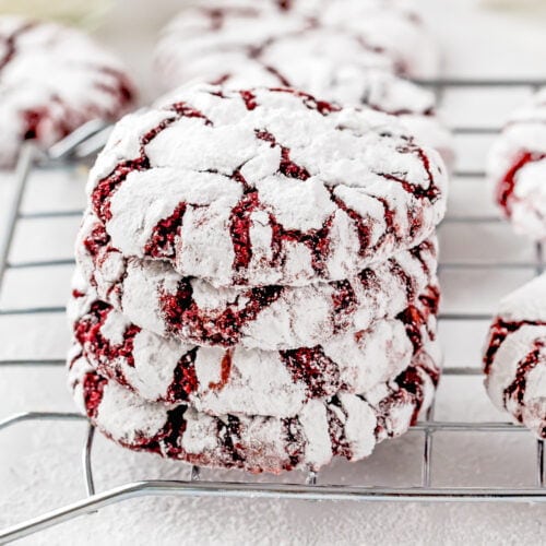 A stack of three eggless red velvet crinkle cookies coated in powdered sugar sits on a cooling rack, with more cookies in the background.