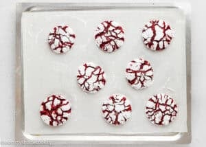 A baking tray with eight eggless red velvet crinkle cookies covered in powdered sugar, arranged on parchment paper.