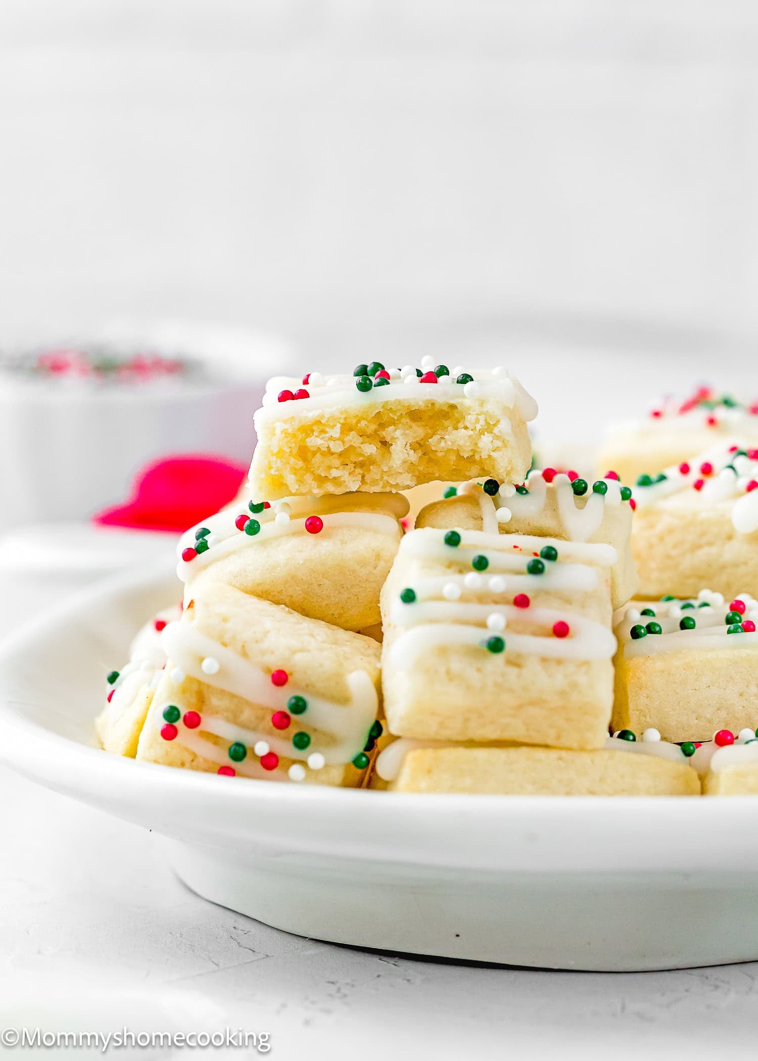 A plate of rectangular Eggless Sugar Cookie Bites topped with white icing and red, green, and white sprinkles, with one cookie showing a bite taken out.