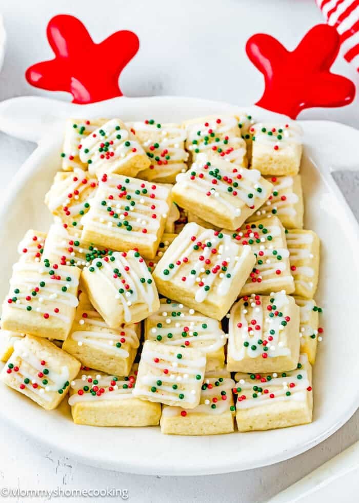 A plate of square Eggless Sugar Cookie Bites topped with white icing and red, green, and white sprinkles, served on a dish with red decorative antlers.