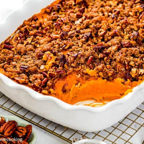 A white baking dish filled with Eggless Sweet Potato Casserole topped with a pecan crumble, with a portion scooped out, sits on a cooling rack.