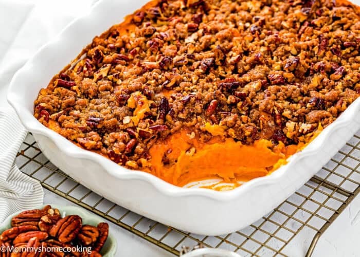 A white baking dish filled with Eggless Sweet Potato Casserole topped with a pecan crumble, with a portion scooped out, sits on a cooling rack.