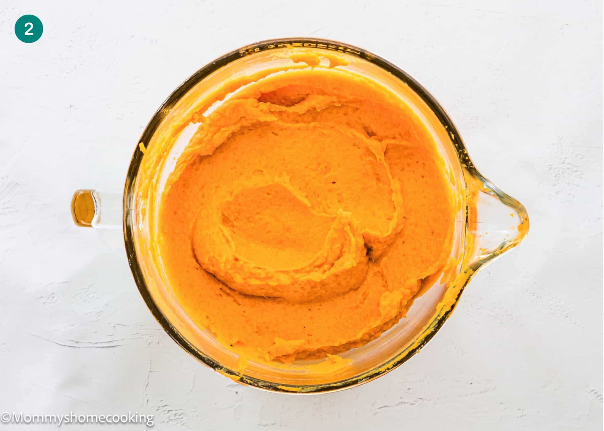 A glass mixing bowl filled with smooth, mashed orange sweet potatoes for an Eggless Sweet Potato Casserole sits on a white surface.