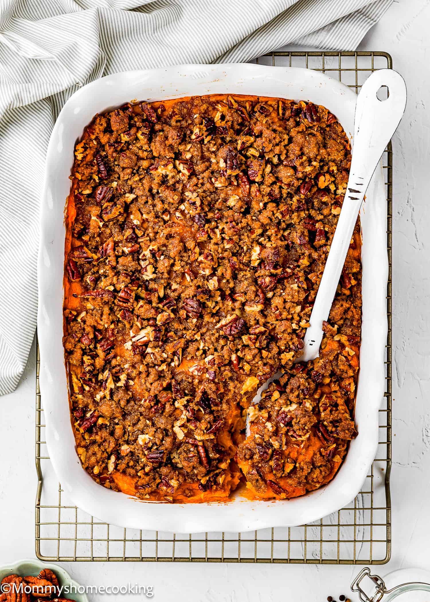 A white baking dish filled with Eggless Sweet Potato Casserole, topped with a pecan and crumb topping, with a serving spoon and a portion already scooped out.