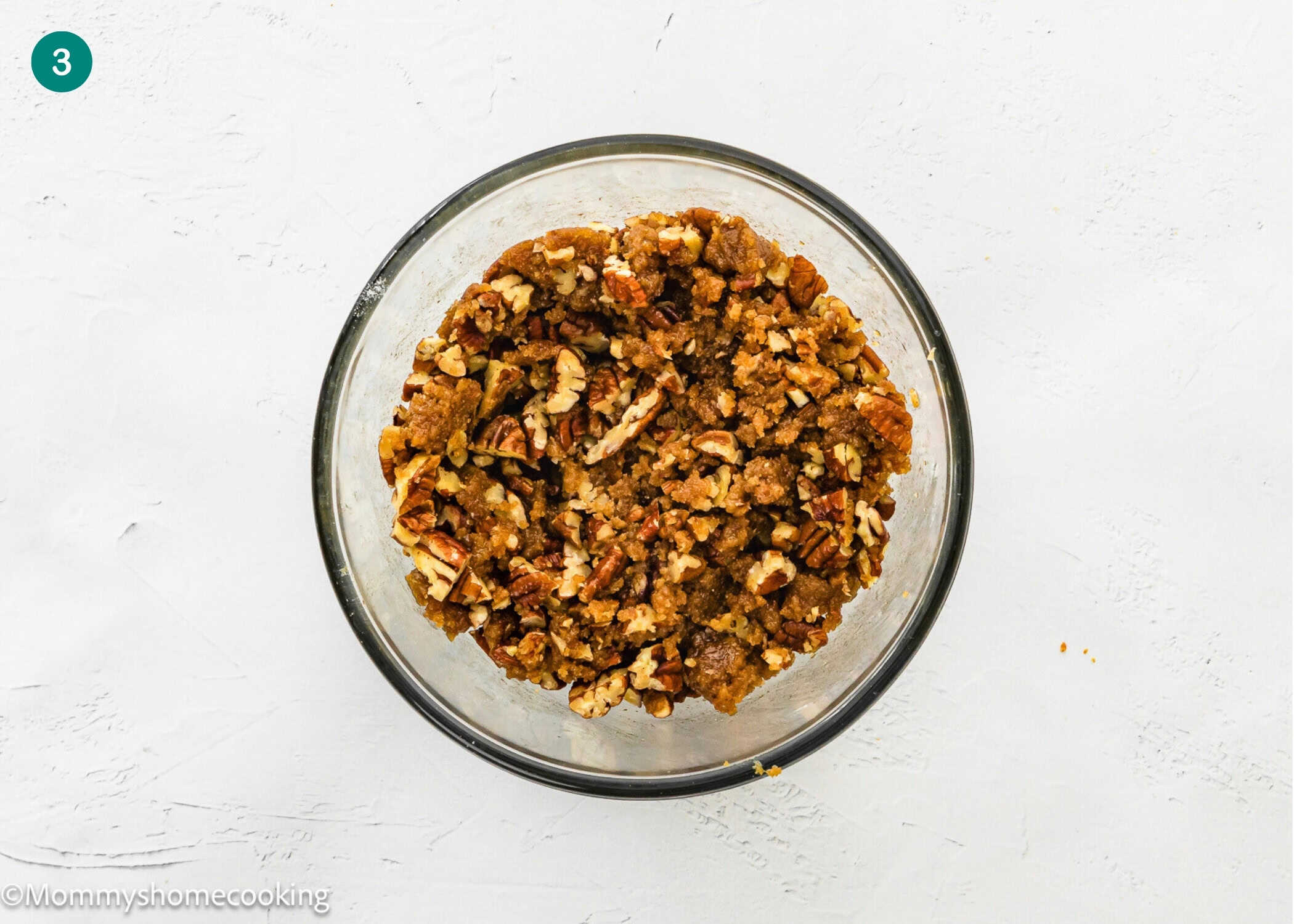 A clear glass bowl filled with a crumbly mixture containing chopped pecans for Eggless Sweet Potato Casserole, placed on a white surface.
