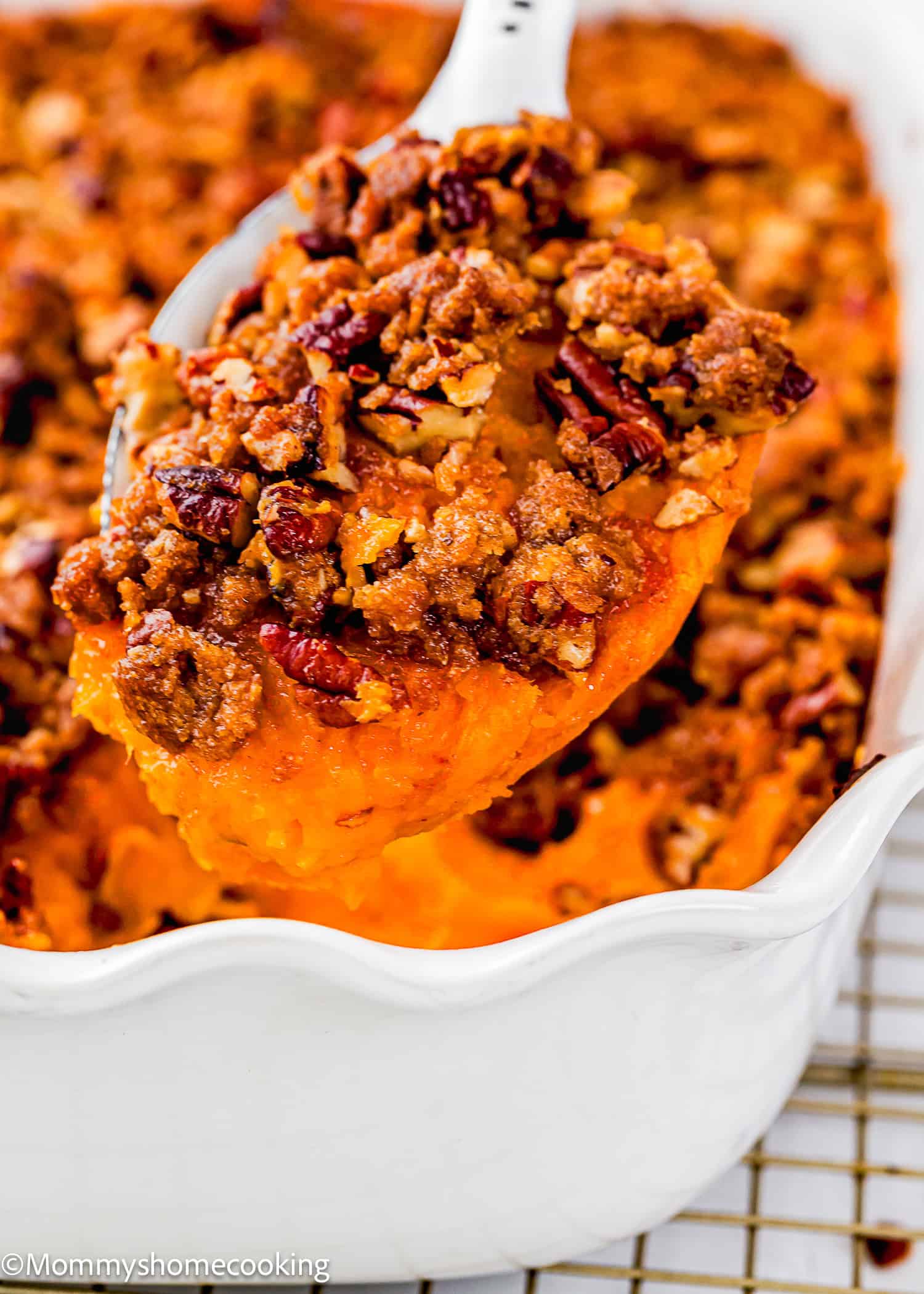 A close-up of a spoon lifting eggless sweet potato casserole with a crumbly pecan topping from a white baking dish.