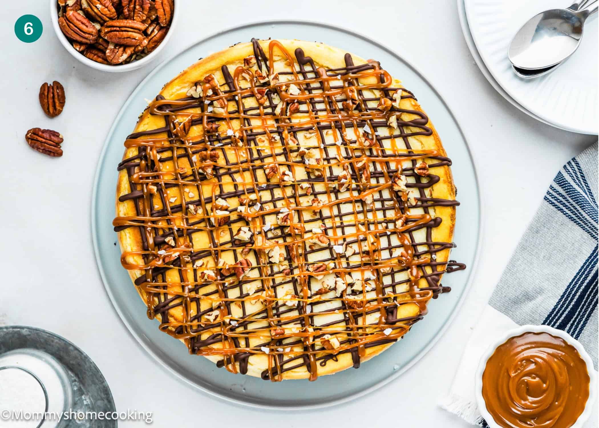 An Eggless Turtle Cheesecake topped with chopped pecans and drizzled with chocolate and caramel sits on a glass plate, surrounded by a bowl of pecans, a dish of caramel, and tableware.