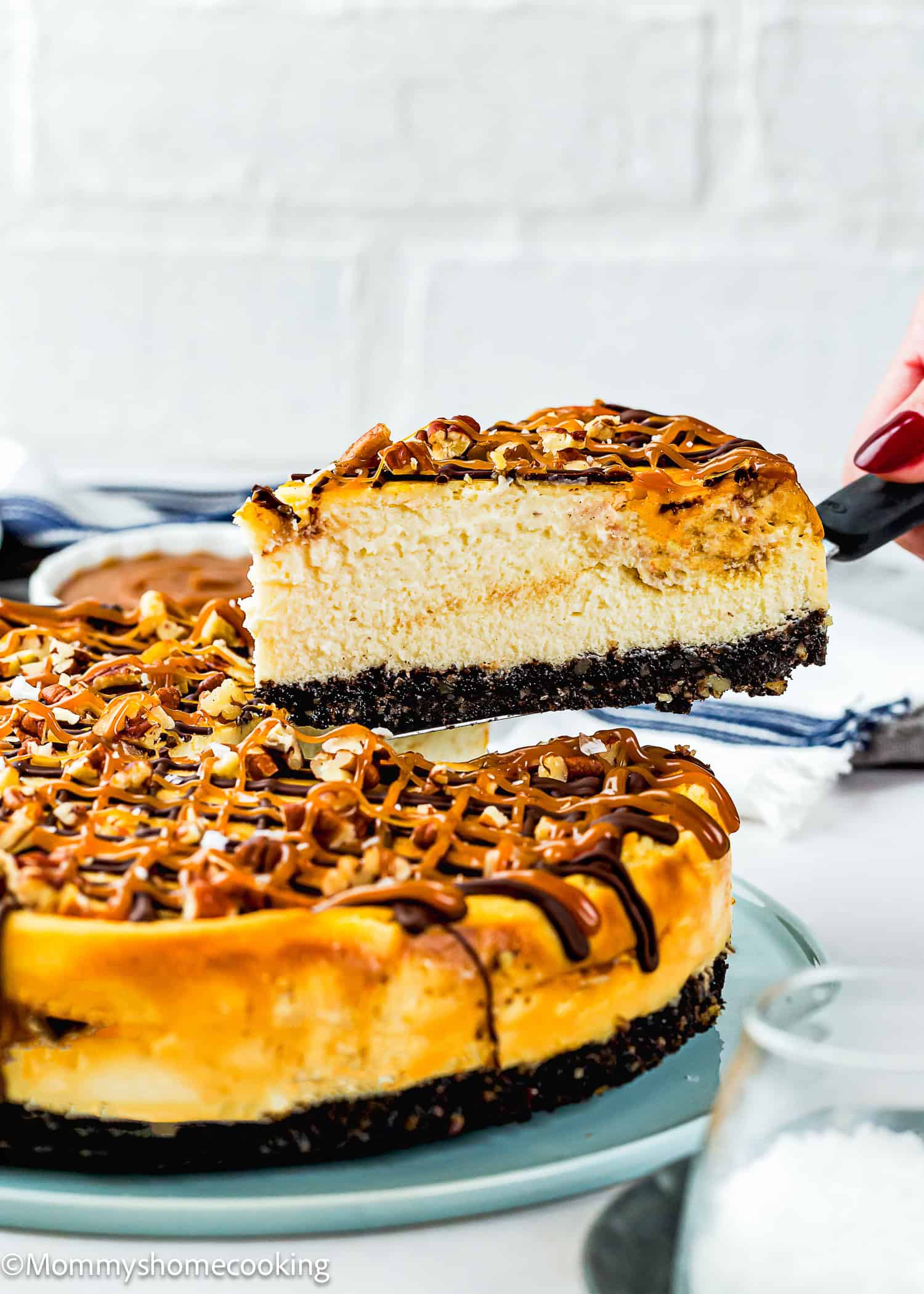 A hand lifts a slice of Eggless Turtle Cheesecake with caramel drizzle and a chocolate cookie crust from a whole cheesecake on a plate.