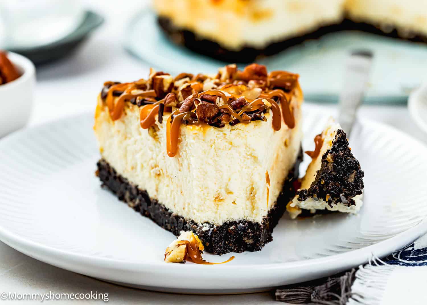 A slice of Eggless Turtle Cheesecake with a dark cookie crust, topped with chopped nuts and caramel drizzle, sits on a white plate with a forkful beside it.