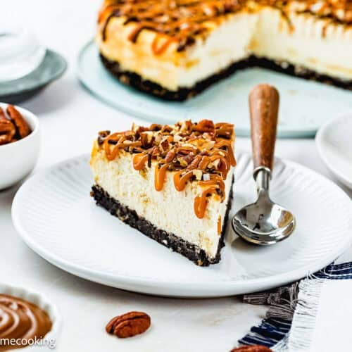 A slice of Eggless Turtle Cheesecake with caramel drizzle and chopped pecans sits on a white plate with a spoon; the rest of the cheesecake is in the background, surrounded by pecans and caramel sauce.