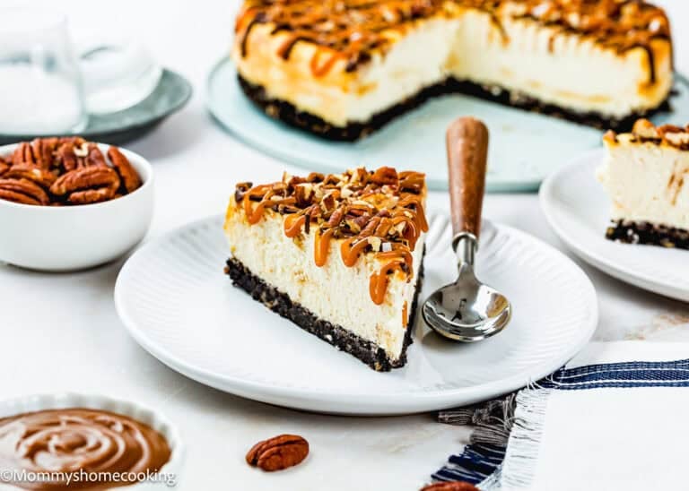 Slices of eggless turtle cheesecake with caramel drizzle and chopped pecans are placed on a white plate with a spoon. The rest of the cheesecake is in the background, surrounded by pecans and caramel sauce.