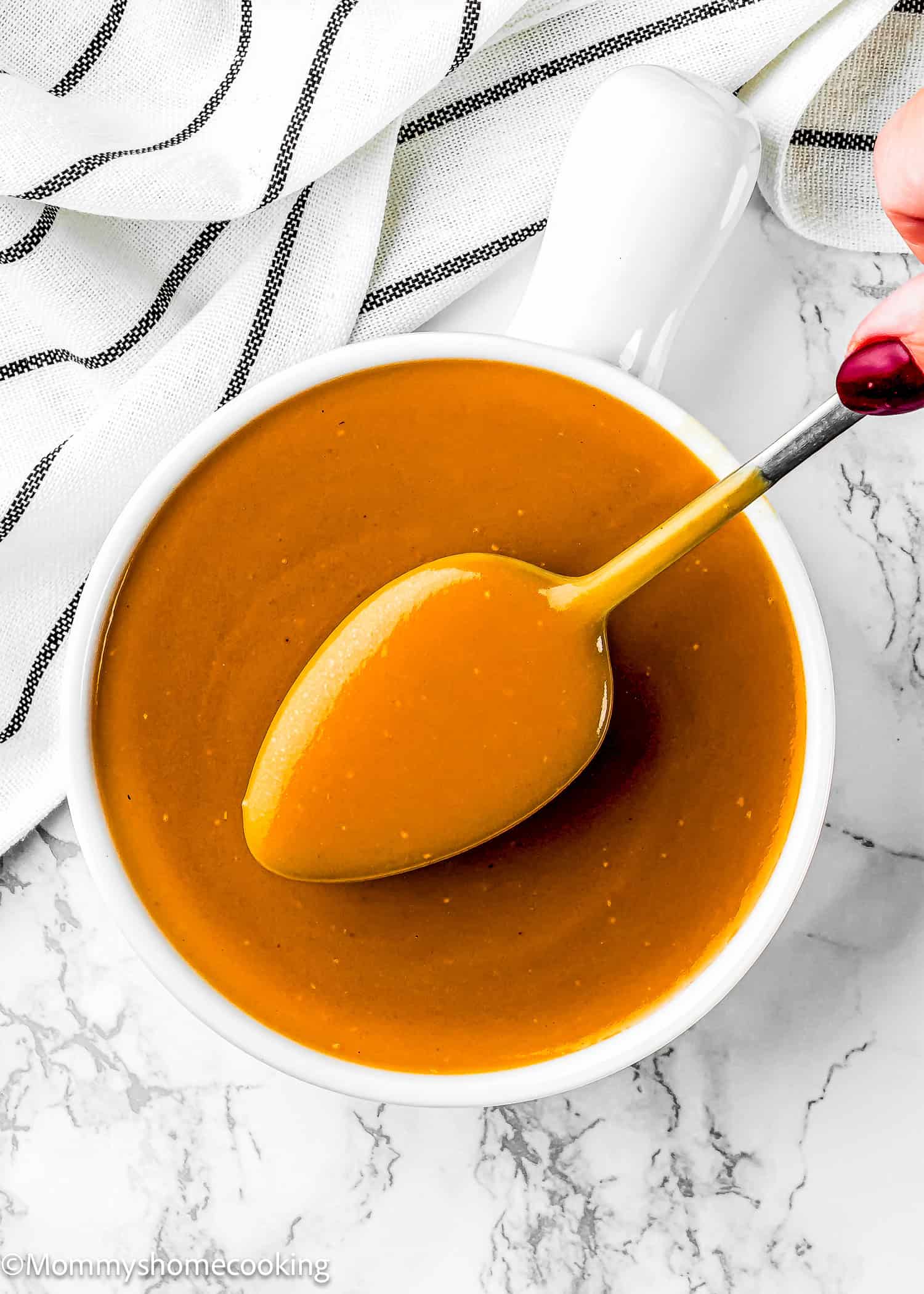 A spoon is held above a white bowl filled with smooth, flavorful gravy on a marble surface, alongside a striped cloth. Try this no dripping gravy recipe for rich taste.